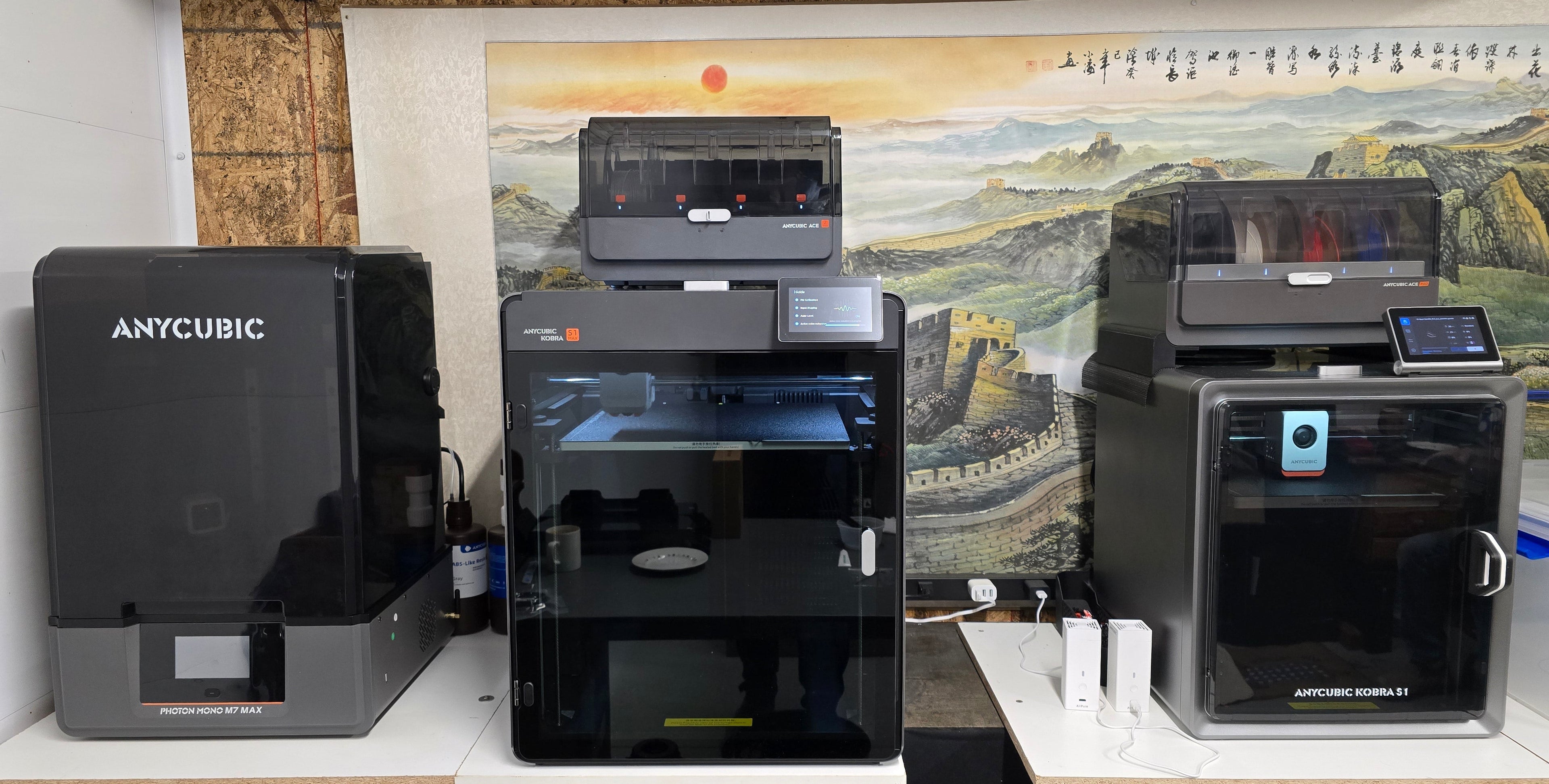 Three 3D printers on a shelf with a scenic wall art in the background
